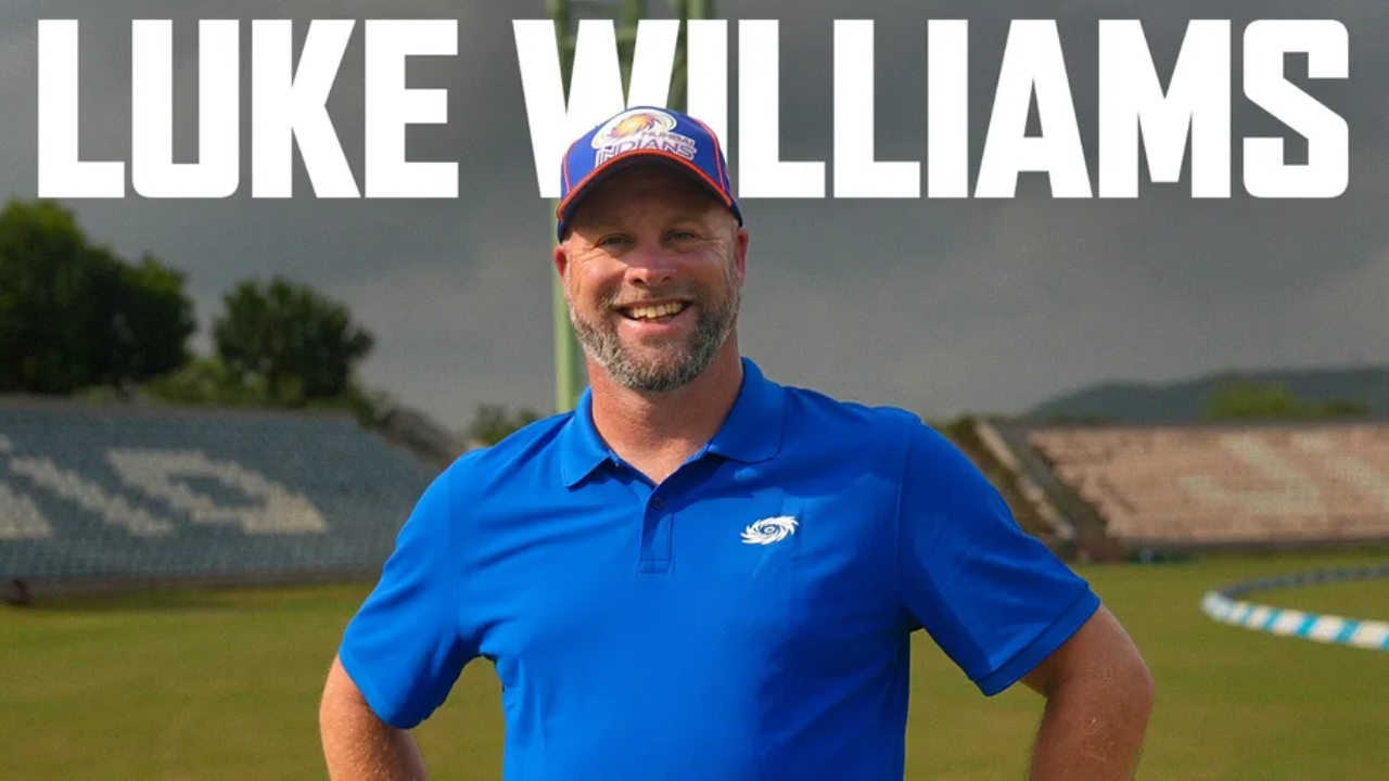 Luke Williams, cricket coach, smiles while wearing a blue Mumbai Indians polo shirt and cap at a cricket stadium.