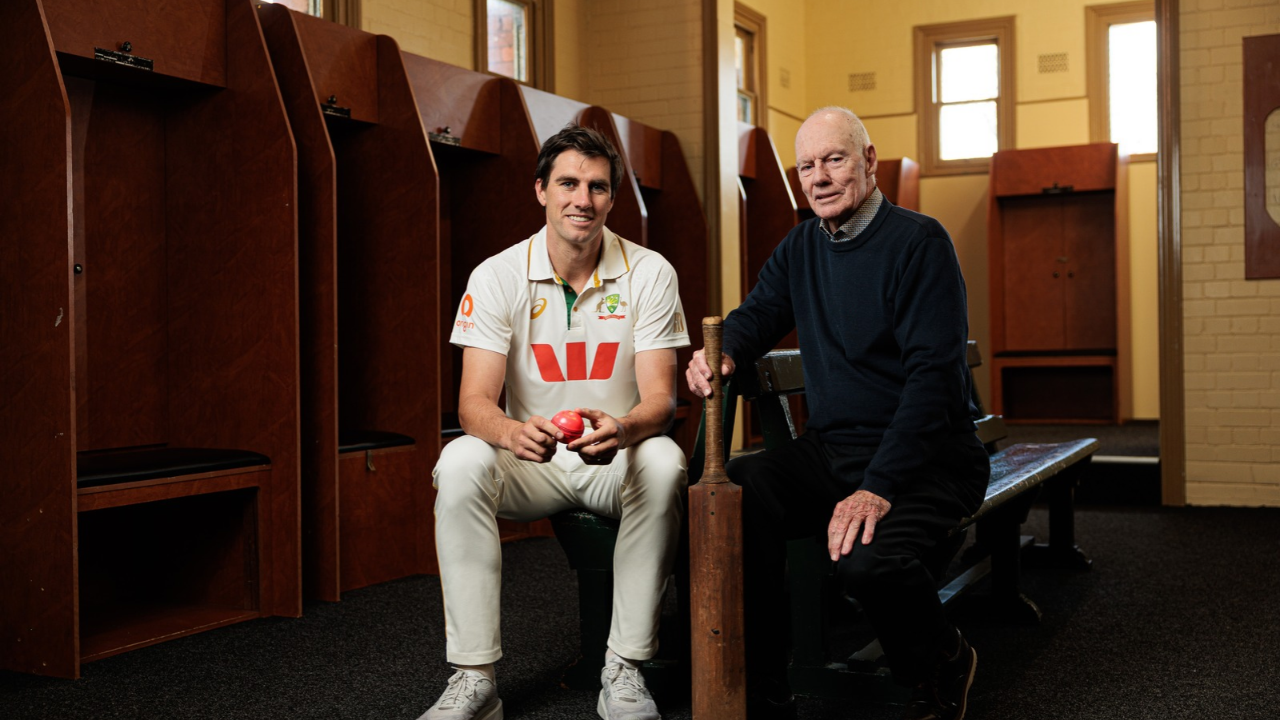 Australian cricket captain Pat Cummins and former cricketer Greg Chappell sitting together in a cricket dressing room, holding a cricket ball and a wooden bat.