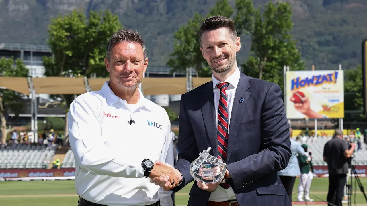 ICC umpire Richard Kettleborough, wearing a white officiating shirt, shakes hands with a suited official holding a glass award on a cricket ground.