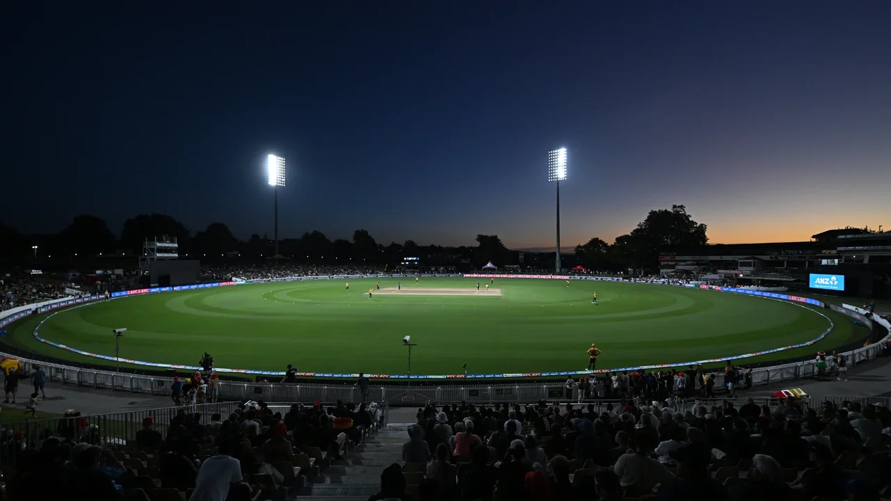 Panoramic view of Seddon Park cricket stadium in Hamilton, New Zealand, during a floodlit evening cricket match with spectators in the stands.