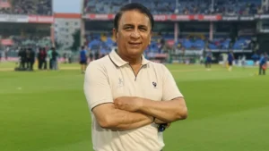 Sunil Gavaskar, former Indian cricket captain and batting legend, stands with his arms crossed on a cricket ground with stadium stands in the background.